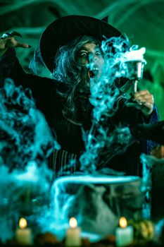 Portrait of witch with awfully face in creepy surroundings and smoky green background drinks magic potion from the goblet.