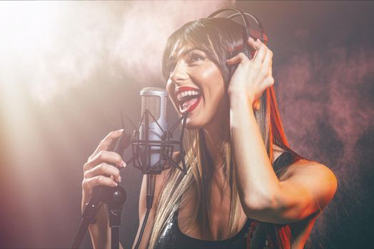 A young woman singer with headphones in front of the microphone. Sing with mouth wide open and with an expression of happiness on her face.