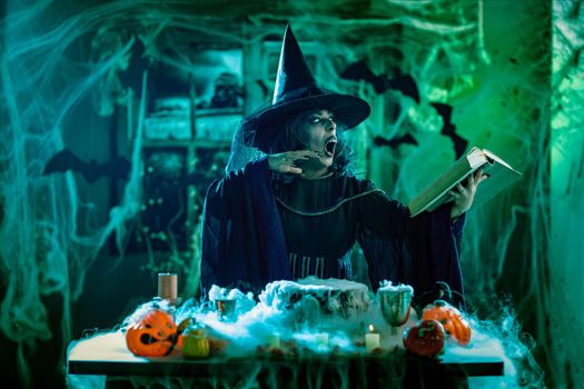 Witch with awfully face in creepy smoky green surroundings full of cobweb is reading recipe of magic drink. Halloween concept.