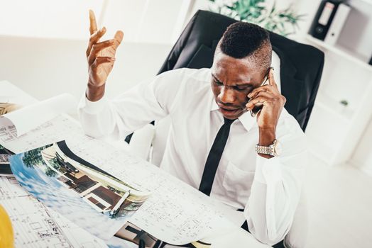 Pensive African architect talking on smartphone and analyzing blueprint.