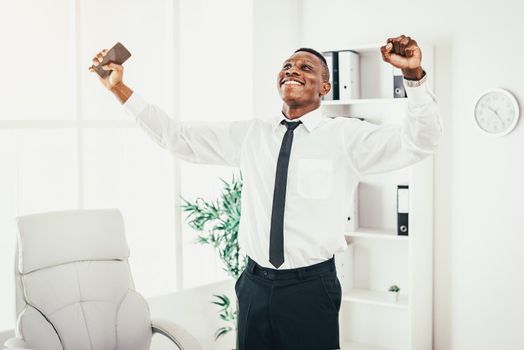 Successful African businessman celebrating success with raised arms in the modern office.