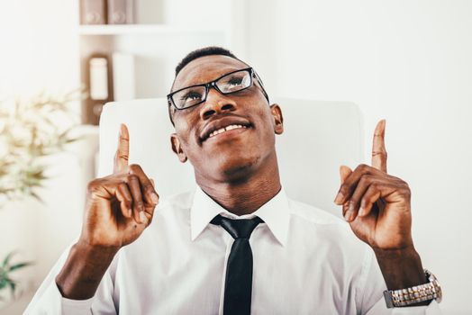 African businessman is sitting in the office, satisfied with success and shows fingers up.