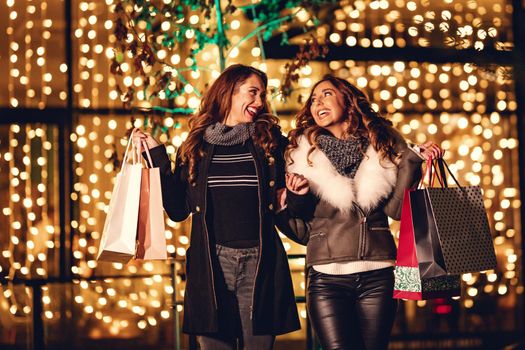Two female friends enjoy the night in shopping, they are looking each other, and laughing with shopping bags in their hands.