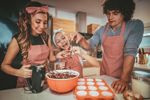 Happy parents and their daughter are preparing cookies together in the kitchen. Little girl helps to her parents with mixer.