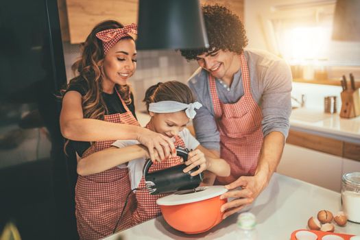 Happy parents and their daughter are preparing cookies together in the kitchen. Little girl helps to her parents mixing dough with mixer.