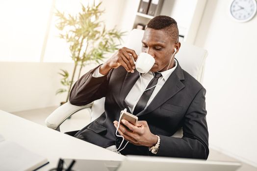 African businessman is sitting in the office, drinking coffee and listening  music in headphones.