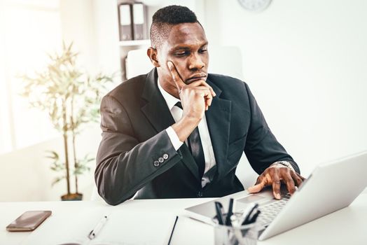 Pensive African businessman working on laptop in modern office.