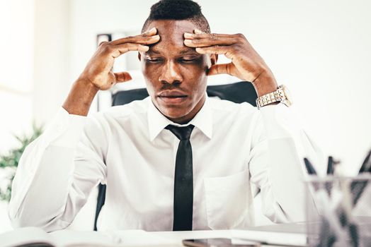 Anxious African businessman sits in the office and holds his head with his hands.