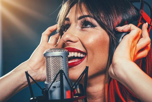 Portrait of a young woman singer with headphones in front of the microphone. Sing with mouth wide open and with an expression of happiness on her face.