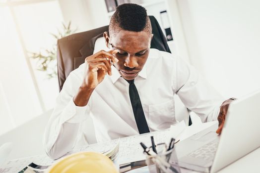 Pensive African architect analyzing blueprint and working on laptop in modern office and planning what to do next.