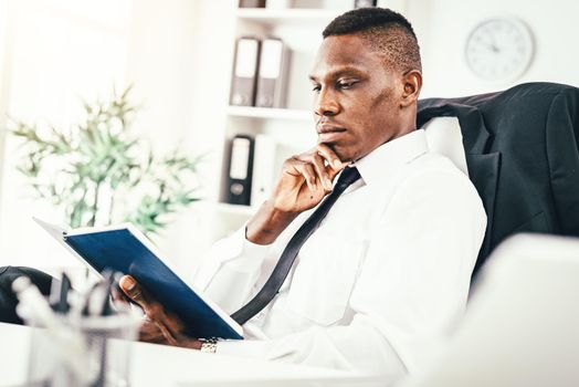 Pensive African businessman working in modern office and reading his notes in notebook and planning what to do next.