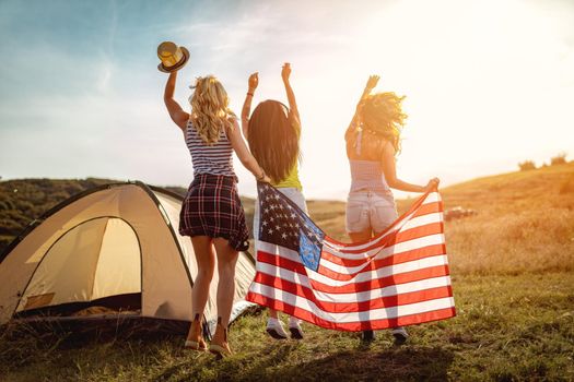 Happy young female friends enjoys a sunny day in nature. They're dancing and holding an american flag in front a campsite tent. Rear view.