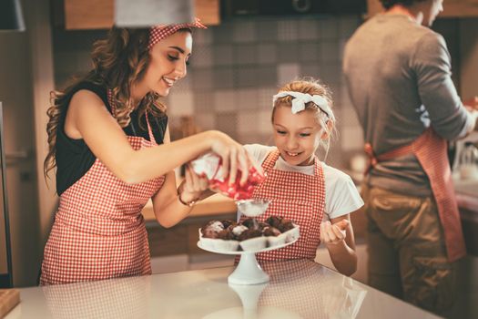 Mom and daughter are sprinkling powdered sugar on roasted chocolade muffin.