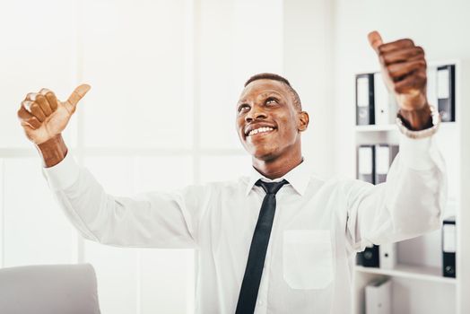 Successful African businessman celebrating success with raised arms and thumbs up in the modern office.