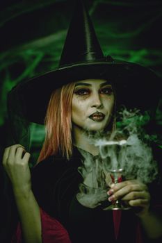 Witch with awfully face in creepy surroundings and smoky green background drinks magic potion from the goblet.