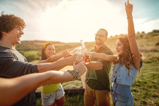 Happy young friends enjoy a sunny day in nature. They're clinking beer bottles, laughing and talking happy to be together.