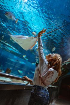 Young girl standing outstretched against aquarium glass fascinated by ocean world and touches the fishes in an oceanarium tunnel.