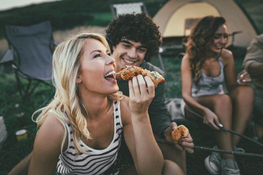 Happy young couple enjoy a sunny day with friends at the mountain. They're laughing and eating grilled sausages near tent.