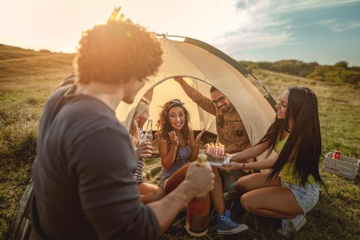 Young people have a good time in camp in nature. They're celebrating a birthday, laughing and greeting to their friend with birthday cake, happy to be together.