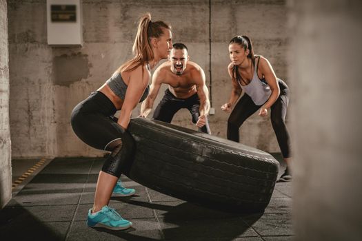 Young muscular woman flipping a tire on cross fit training at the gym and man and woman are looking at her.