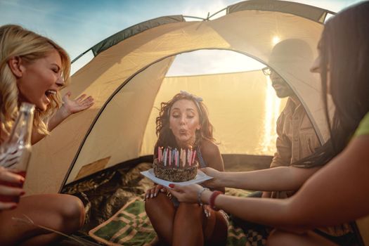 Young people have a good time in camp in nature. They're celebrating a birthday, laughing and greeting to their friend with birthday cake, happy to be together.