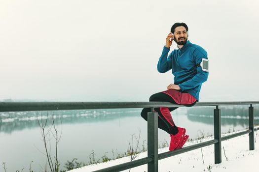 A male runner with headphones on his ears taking a break in the public place during the winter training outside beside the river. Copy space.