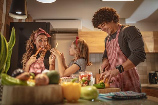 Cute little girl and her beautiful parents are cutting vegetables and smiling while making pickle in kitchen at home