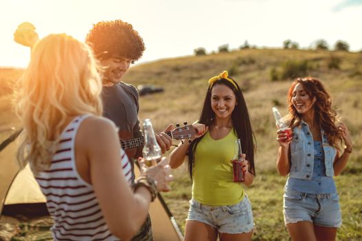 Young people have a good time in camp in nature. They're resting, laughing and singing with music from ukulele, happy to be together.