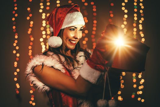 Beautiful young smiling woman in Santa Claus costume holding red present and having fun looking what is in it..