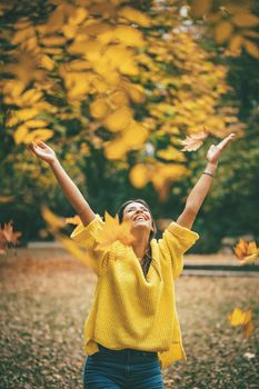 Beautiful young woman enjoying in sunny forest in autumn colors. She is holding golden leaves and standing under the tree. 