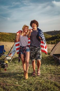 Happy young couple enjoys a sunny day in nature. They're hugging each other, wrapped in american flag.