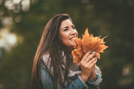 Cute young woman enjoying in sunny forest in autumn colors. She is holding golden yellow leaves.