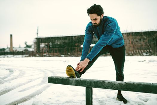 Active young  man stretching and doing exercises in the public place during the winter training outside in while it snowing. Copy space. 