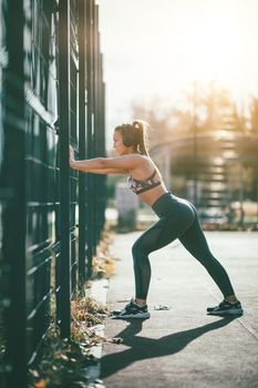 Young female runner, with headphones on her ears, doing stretching exercise on metal fence, preparing for morning workout.