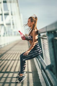 Young fitness woman is resting after hard training on the river bridge and drinking water.