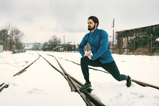 Active young man stretching and doing exercises in the public place among old railroad during the winter training outside in. Copy space. 