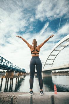 Young fitness woman standing by river bridge with open arms and preparing for morning workout with bottle of  water.