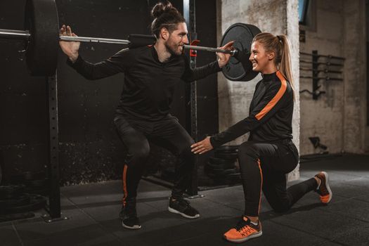 Young muscular man doing squat exercise with barbell at the gym with support of his personal female trainer. 