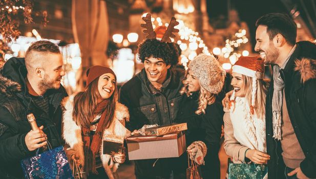 Six smiling young friends standing outside and holding many shopping paper bags, boxes and champagne bottle in city night.