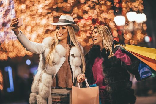 Cheerful young female friends are having fun in city street at the Christmas night. They are taking selfie with a smartphone and talking something funny.