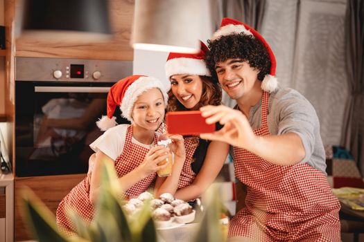 Happy parents and their daughter with santa's cap are preparing meal together in the kitchen. They are taking selfie with smartphone.