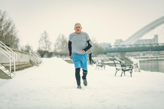 Active senior man running and doing exercises by the river during the winter training outside in. Copy space.