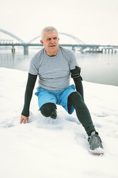 Active senior man stretching and doing exercises by the river during the winter training outside in.