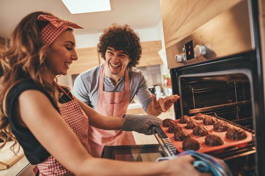 Young happy couple pulling baked muffins from the oven.