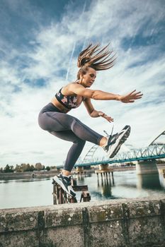 Beautiful young woman jumping after successful training on the wall by the river bridge.
