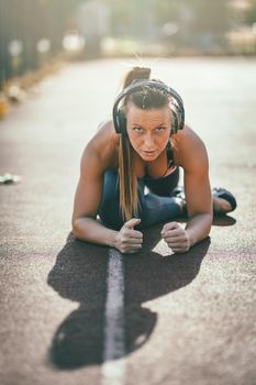 Young female runner, with headphones on her ears, doing stretching exercise on a public place, preparing for morning workout.