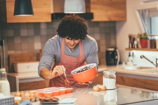 Young handsome man making cookies in his kitchen for his friends. He is pouring chocolate dough into molds.