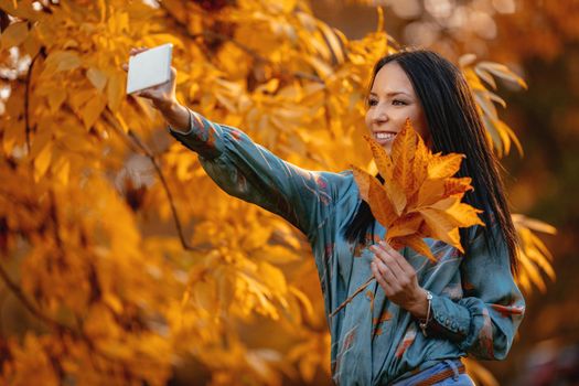 Modern cute girl taking a selfie in park in autumn with dry leaves in her hand.  