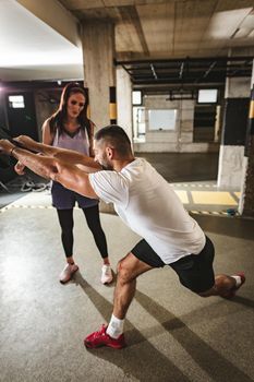 Young handsome woman having workout with a suspension straps at the gym supported of her personal male trainer. 