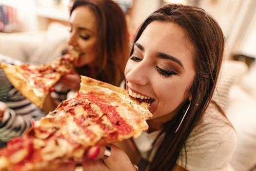 Excited young women are eating pizza sitting on sofa. They have great weekend in nice company indoors. 